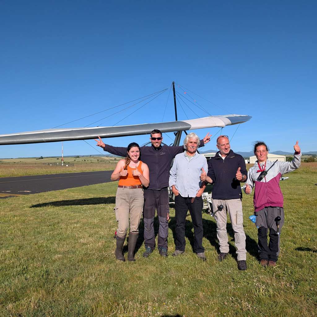 Five people stand on a grassy airfield in front of a glider, smiling and giving thumbs up under a clear blue sky.