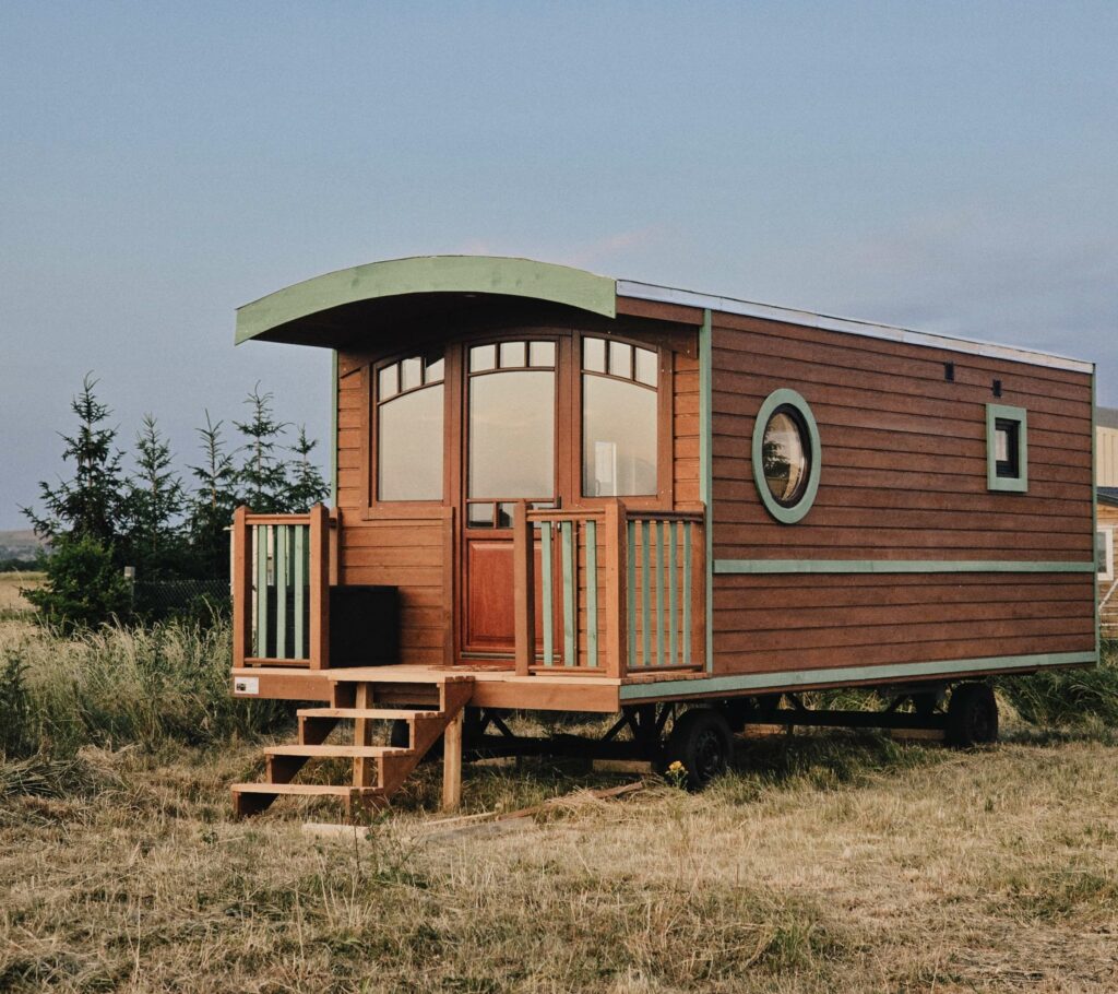 Tiny wooden tiny house on a trailer with curved green roof and arched windows, set in a grassy field with trees nearby.